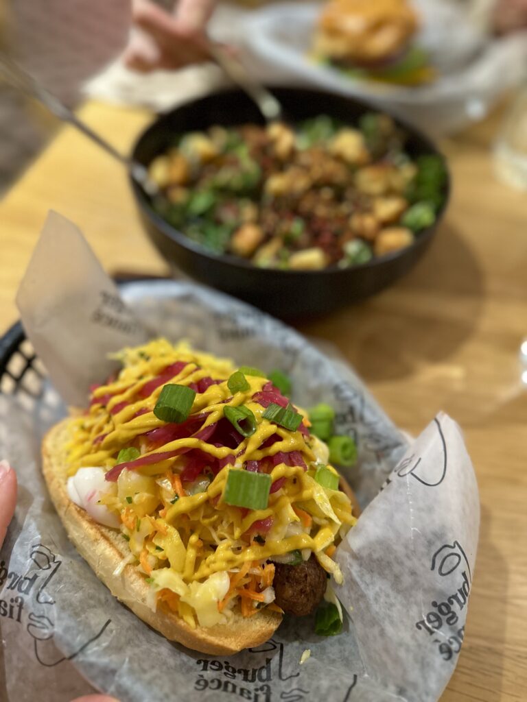 vegan hot dog loaded with coleslaw, green onions, pickled red onions and mustard. kale caesar salad in the background.