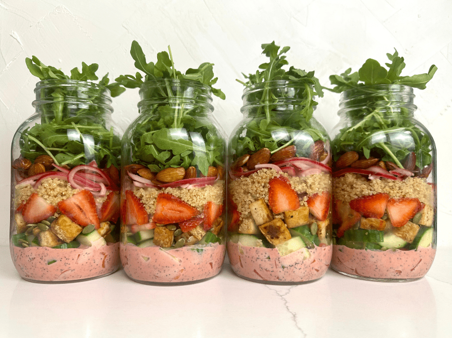 4 salad jar meal preps in a row layered with strawberry poppyseed dressing, bell pepper, cucumber, tofu, seeds, strawberries, quinoa, pickled red onion, and arugula