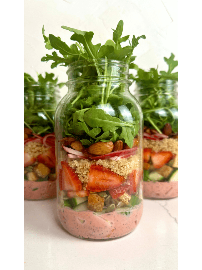 a beautifully layered salad jar with a vibrant pink dressing, fresh sliced strawberries and bright greens sprouting out the top. 