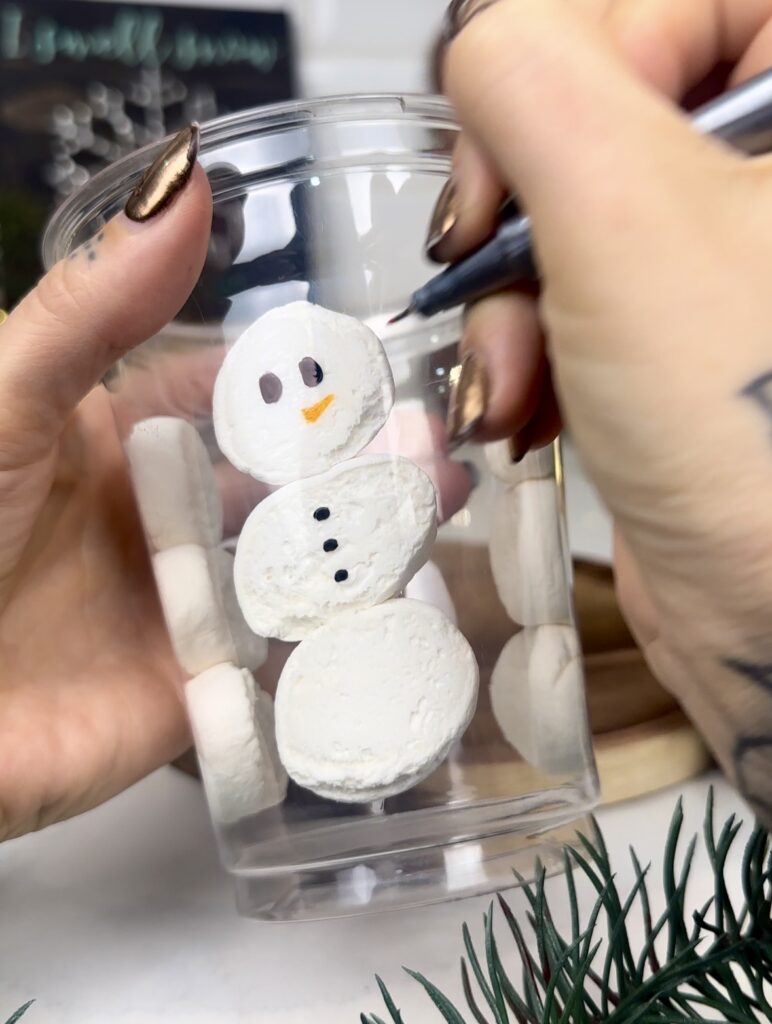 using a sharpie marker to add the eyes, buttons and nose to the snowman drawn on the outside of the cup