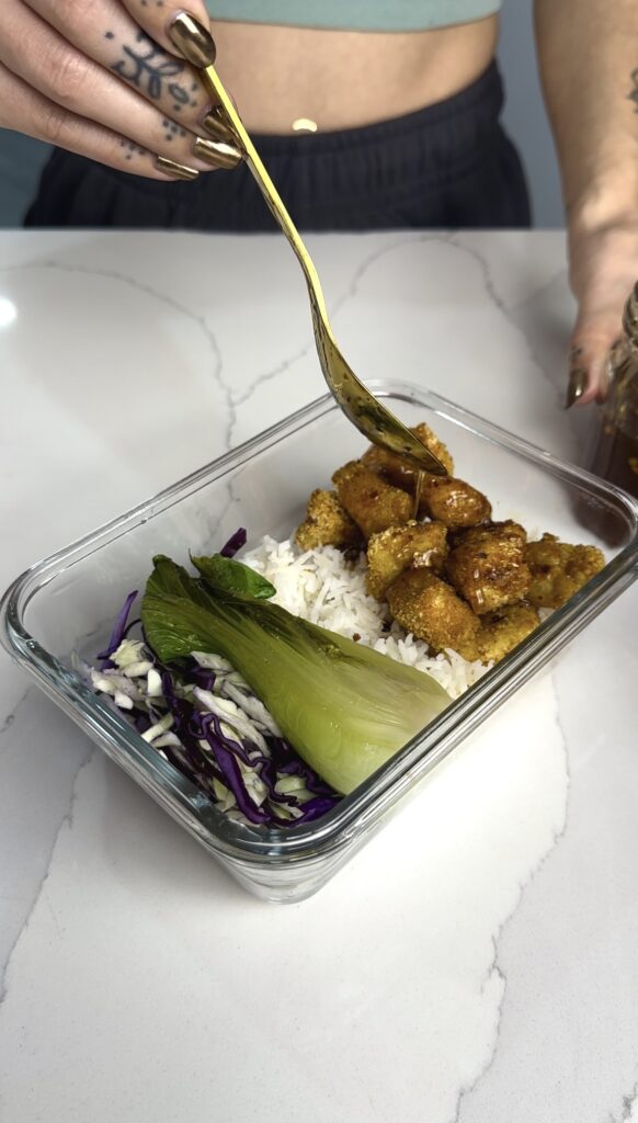 vegan hot and honey sauce being added to the meal prep containers with crispy tofu, bok choy, coleslaw and rice.