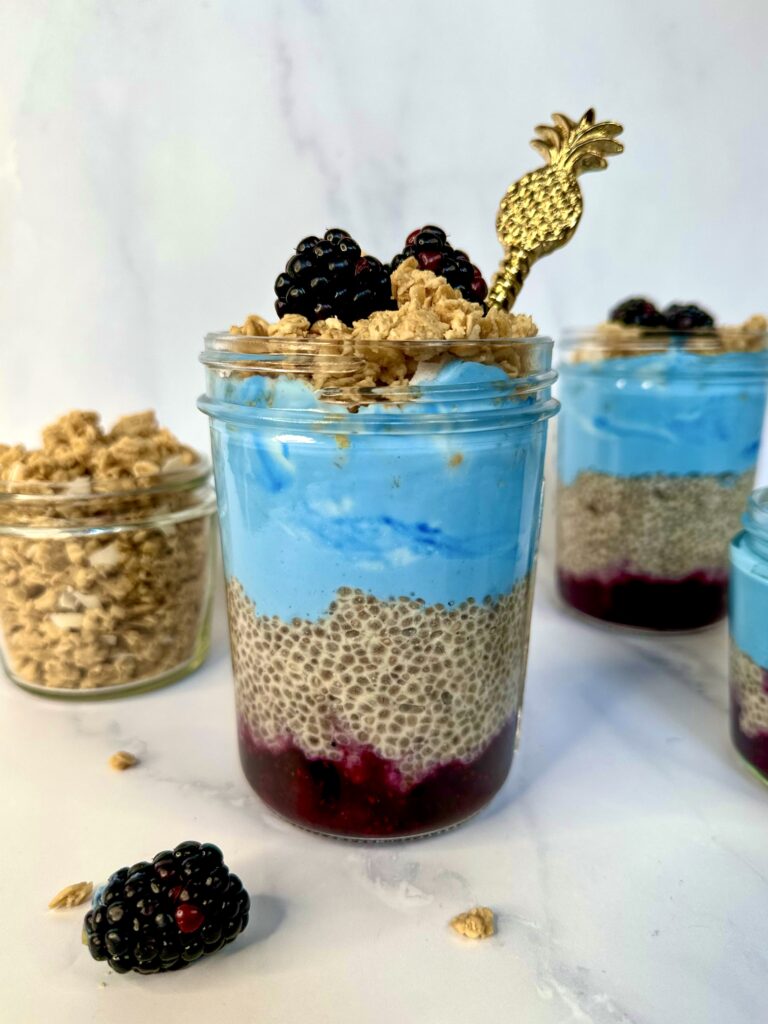 A glass jar with Blueberry Chia Pudding Parfait in it. A layer of jam at the bottom, chia pudding, blueberry yogurt, granola, and fresh berries.