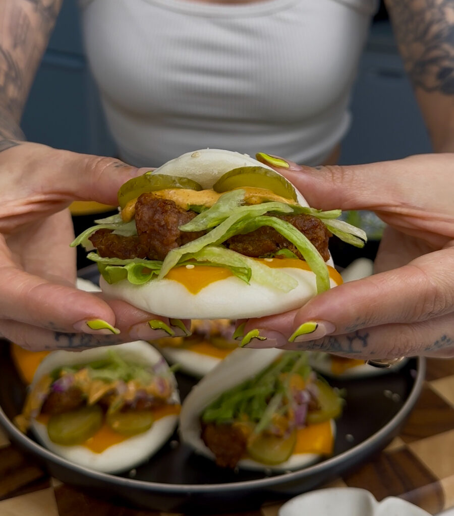 hands holding vegan big mac bao buns with a cheddar slice, beyond beef chunks, lettuce, pickles, and red onion