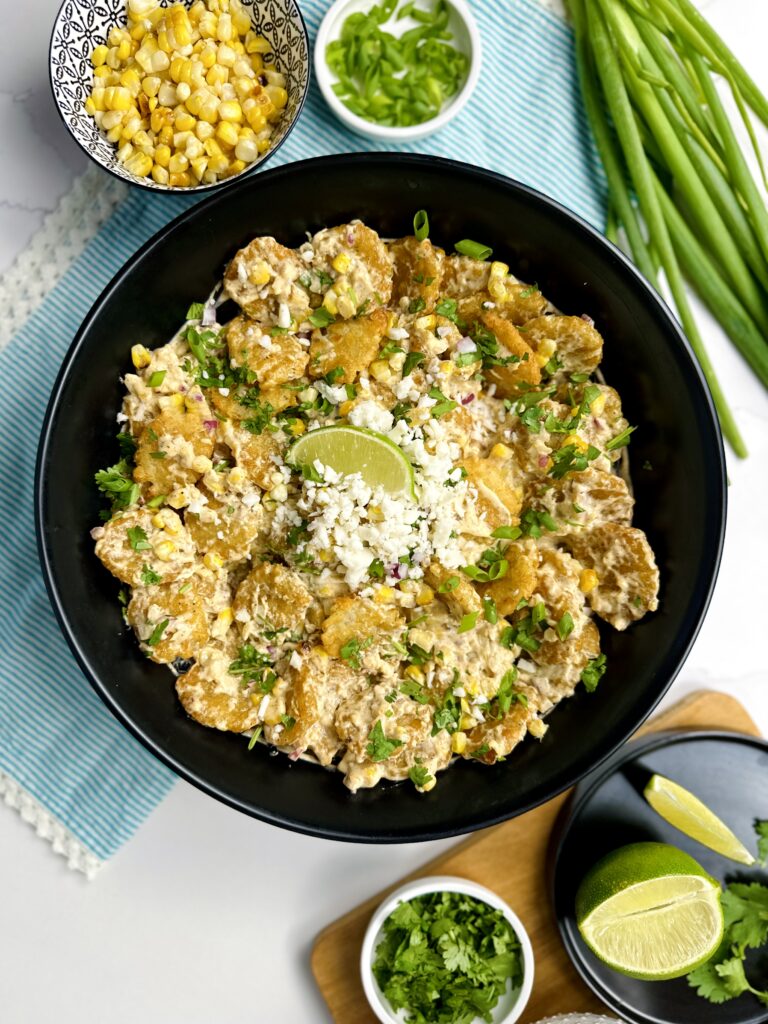 place setting with large bowl of tater tot sweet corn salad, surrounded by corn, green onion, lime, cilantro and vegan feta cheese