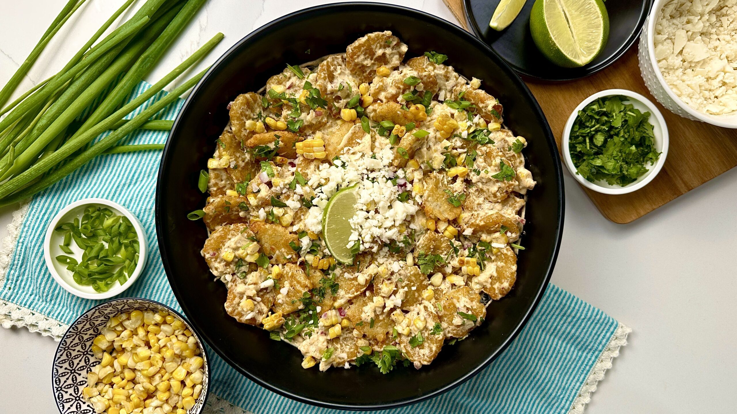 place setting with large bowl of tater tot sweet corn salad, surrounded by corn, green onion, lime, cilantro and vegan feta cheese