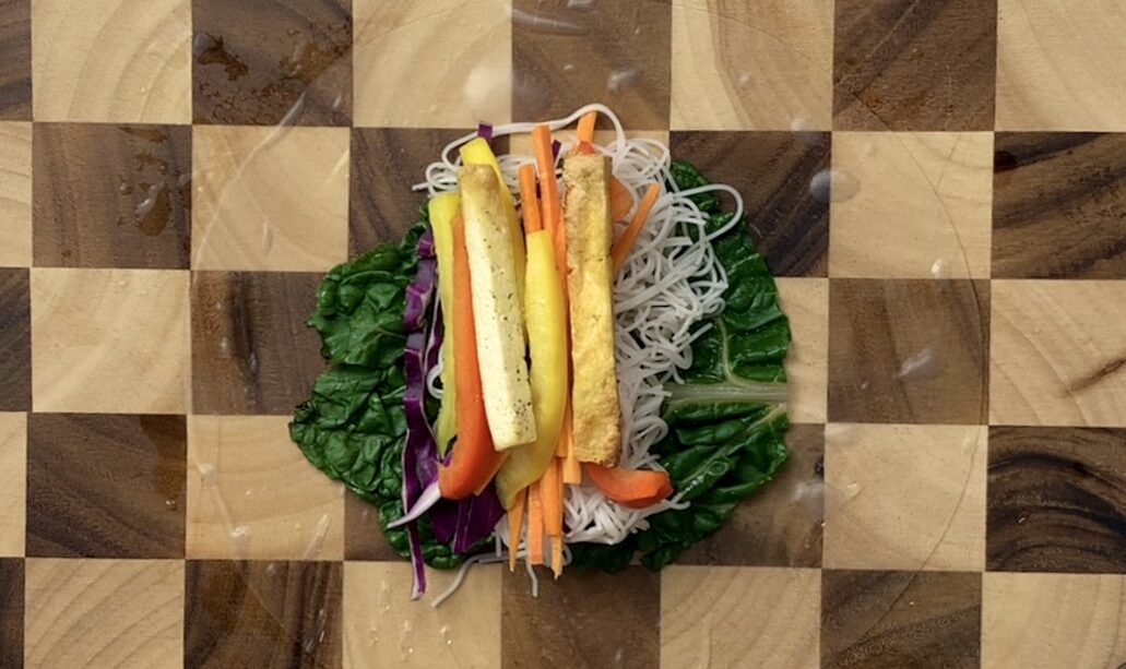 wooden checkered cutting board with rice paper, Swiss chard, vermicelli noodles, cabbage, peppers and crispy tofu sticks